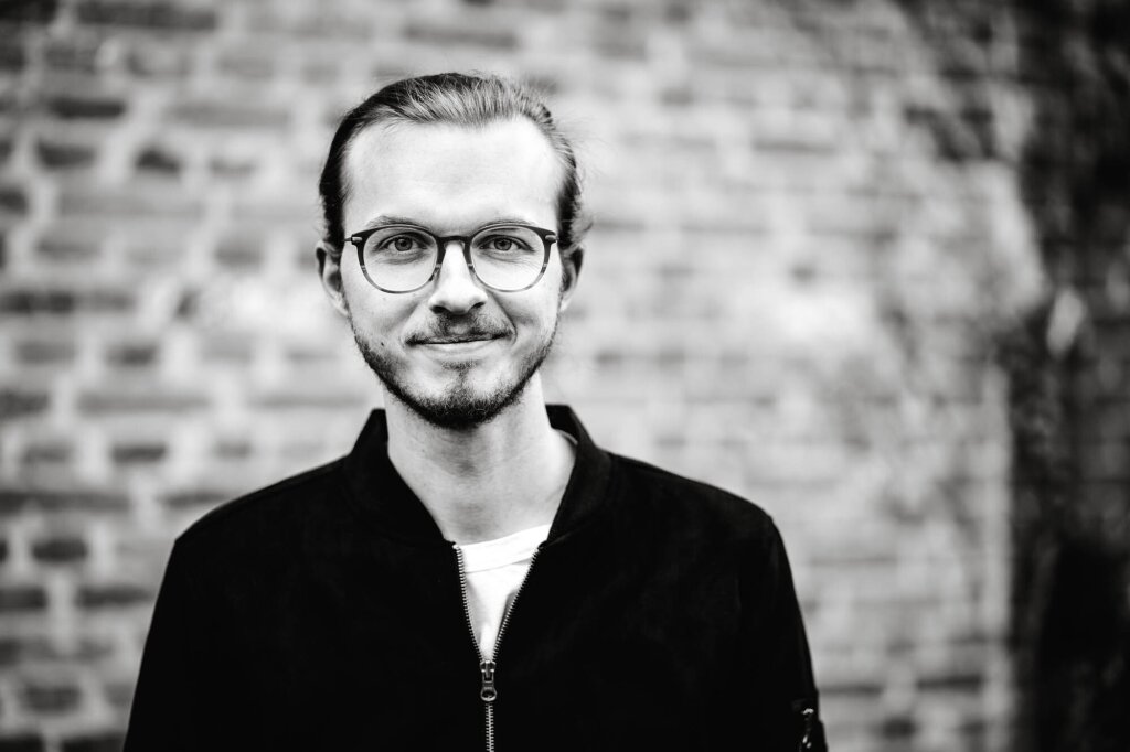 Portraitfoto von Webentwickler Johannes Schuba. Er trägt Brille, lacht leicht in die Kamera und trägt seine Haare in einem Dutt.